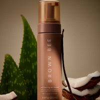The self-tanning mousse bottle styled beside an aloe plant and a halved coconut