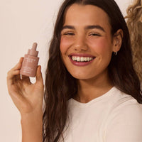 A smiling woman in a white t-shirt holding the H7 face tanning serum