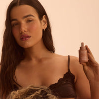A dark-haired woman holding the H7 face tanning serum, shown with an even facial glow