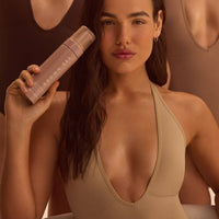A dark-haired woman in a tan bodysuit holding the self-tanning mousse, showing an even all-over glow